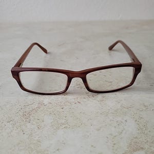 Może przedstawiać: Prostokątne okulary z ciemnobrązową oprawką o fakturze drewna. Przezroczyste soczewki są osadzone w oprawce, a zauszniki sięgają do uszu. Okulary są umieszczone na neutralnym tle.