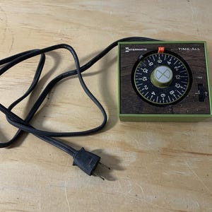 May include: A vintage Intermatic Time-All timer with a black dial and green and brown accents. The timer has a black power cord and plug. The dial is marked with numbers and a green knob. The text "TIME-ALL" is visible.