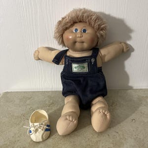 Op de afbeelding: Een vintage Cabbage Patch Kid-pop met blond, pluizig haar en blauwe ogen, gekleed in een spijkerbroek. Een paar witte sneakers met blauwe strepen liggen naast de pop. De pop zit met uitgestrekte armen.