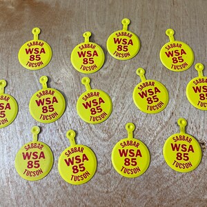 May include: A collection of yellow, circular tags with a loop at the top. Each tag features the text "SABBAR WSA 85 TUCSON" in red. One tag has the word "ELDORADO" and a signature. The tags are scattered on a wooden surface.