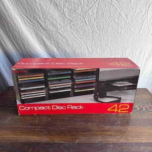 May include: A rectangular cardboard box for a Radio Shack Compact Disc Rack 42. The box features a red and black design with a graphic showing a black CD storage rack filled with compact discs, a CD player, and headphones. The text reads Compact Disc Rack 42.