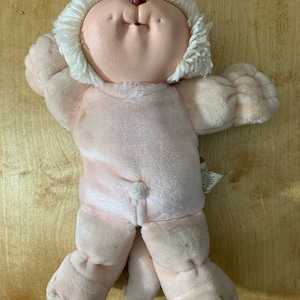 May include: A vintage Cabbage Patch Kids doll with a light pink body and a white yarn hairstyle. The doll has a round face with blue eyes and a small nose. The doll is lying on a wooden surface.