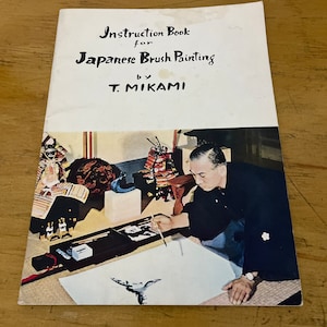 以下が含まれることがあります： T.ミカミ著の日本の筆絵の指導書。表紙には、筆で絵を描く男性の写真が写っており、背景には日本の美術品や装飾品が写っています。本は木製の表面に開かれています。