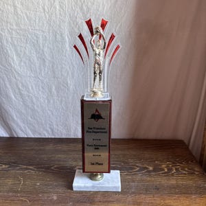 Puede incluir: Un trofeo con una figura de jugador de baloncesto, detalles rojos y transparentes, y una base de mármol. El trofeo tiene una placa rectangular con el texto "San Francisco Fire Department" y "1st Place".