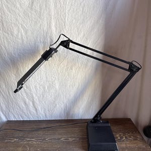 May include: A black adjustable desk lamp with a rectangular base and a long, articulated arm. The lamp's head is angled downward, and the design is minimalist and functional. The lamp is on a wooden surface.