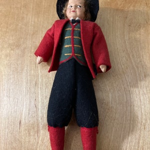 Op de afbeelding: Een vintage pop gekleed in traditionele klederdracht. De pop draagt een zwarte hoed, een rode jas, een groen en goud vest, een zwarte broek, rode sokken en zwarte schoenen. De pop heeft een geschilderd gezicht en staat rechtop.