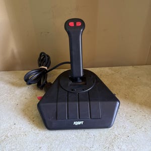 May include: A black gaming joystick with a vertical handle featuring two red buttons at the top. The base is triangular with a black cord and the word KRAFT printed in white on the front. The device has a smooth plastic finish.
