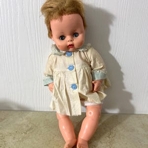Puede incluir: Muñeca de bebé vintage con cabello rubio, ojos azules y un vestido crema con botones azules. La muñeca está sentada. Un juguete clásico para coleccionistas, un objeto de colección nostálgico.