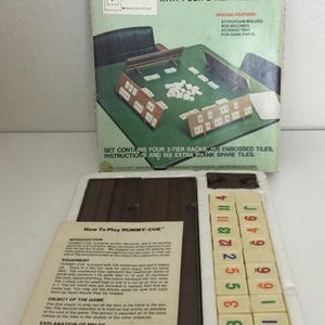 May include: A vintage Rummy-Cue Tile Rummy Game box set. The box is green, red, and white, with the game title prominently displayed. The set includes tiles, racks, and instructions. The game is designed for multiple players.