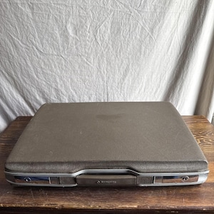 May include: A vintage brown Samsonite briefcase with silver-toned accents. The rectangular case is closed and sits on a wooden surface. The handle is brown and the case has a textured surface. The Samsonite logo is visible on the handle.