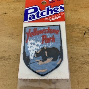 May include: Iron-on patch featuring a blue and white shield with the text "Yellowstone Park" in red. The shield has a black bear and a geyser in the background. The patch is packaged in a clear plastic bag with a red and blue header that reads "Patches" and "Made in U.S.A. by Voyager".