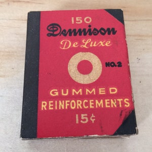 Op de afbeelding: Een rode en zwarte doos met de tekst "150 Dennison De Luxe No.2 GUMMED REINFORCEMENTS 15¢" erop gedrukt.