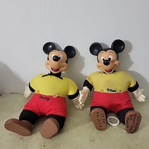 May include: Two vintage Mickey Mouse plush dolls. Each doll has a yellow shirt, red shorts, black shoes, and black ears. One doll is labeled "Dougie" and the other "Stacy". The dolls are sitting on a light-colored surface.