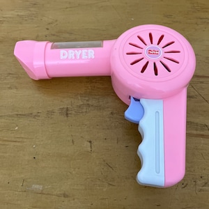 Op de afbeelding: Een roze speelgoed haardroger met het woord "DRYER" aan de zijkant. De handgreep is wit en roze, met een lichtblauwe trekker. De droger heeft een ventilatorachtig ontwerp aan de voorkant en de woorden "My First Buddys" zijn gedrukt.