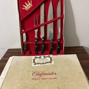 Peut inclure: Un ensemble de couteaux Chefmaster dans une boîte rouge avec un logo de couronne dorée. La boîte comprend également une carte de garantie. Les couteaux ont des manches noirs et sont en acier inoxydable. La boîte est étiquetée "Chefmaster World's Finest Cutlery".
