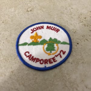 Może przedstawiać: Okrągła naszywka z niebieską obwódką i białym tłem. Naszywka zawiera słowa "JOHN MUIR CAMPOREE '72" w kolorze czerwonym, ze złotym motywem fleur-de-lis i zielonym drzewem.