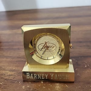 Puede incluir: Un reloj de escritorio dorado con una esfera cuadrada y una base rectangular. La esfera del reloj presenta el nombre "Levitz" y la base está grabada con "Barney Yauch". El reloj tiene un diseño clásico y vintage.