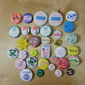 Peut inclure: Une collection de badges vintage avec divers motifs et textes. Les badges comportent des textes tels que "WSA", "Aloha", "Kiss Me" et "Elect Cain". Les couleurs incluent le rouge, le bleu, le jaune et le blanc. Certains badges ont des graphiques.