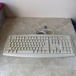 May include: A beige Compaq computer keyboard with a QWERTY layout, a numeric keypad, and a coiled white cable with a purple connector. The keyboard features standard function keys, navigation keys, and the Compaq logo printed in red on the top right.