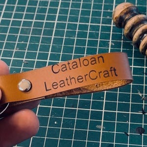 May include: A tan leather keychain featuring a silver metal rivet and a split ring. The leather strap is engraved with the text Cataloan LeatherCraft in a dark font. The keychain is held in a hand, with a wooden burnishing tool visible nearby.