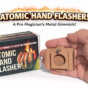 May include: A magic trick prop, the "Atomic Hand Flasher," is shown with its packaging. The box features the text "Atomic Hand Flasher" and an illustration of a hand with flames. A metal gimmick is held in hand.
