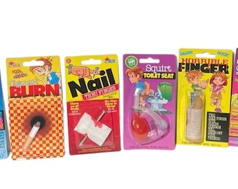 May include: Six different prank novelty items packaged on colorful cards. The items include a squirt lighter, a cigarette burn, a nail through finger, a squirt toilet seat, a horrible finger, and a deluxe squirt ring.