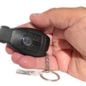 May include: Black car key with silver key ring and chain. The key has several buttons and a circular silver emblem. A small white tag is attached to the key ring.