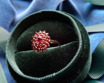 Puede incluir: Un anillo de piedra roja con un engaste en forma de flor, presentado en una caja de anillo de terciopelo verde.