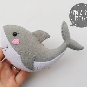 May include: A gray felt shark plush toy with white felt accents and pink felt cheeks. The toy has black embroidered eyes and a stitched mouth. The image includes the text "PDF & SVG PATTERN".