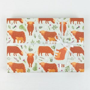 May include: A sheet of wrapping paper with a repeating pattern of brown Highland cows with their calves. The cows are surrounded by green foliage and small pink and purple flowers. The paper is tied with a white ribbon and a gift tag.