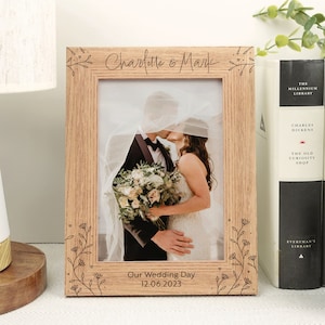 Könnte beinhalten: Ein Holz-Bilderrahmen mit einem floralen Design, das um die Ränder graviert ist. Der Rahmen enthält ein Foto eines sich küssenden Paares mit dem Text "Charlotte & Mark" und "Our Wedding Day 12.06.2023", der unter dem Foto graviert ist.