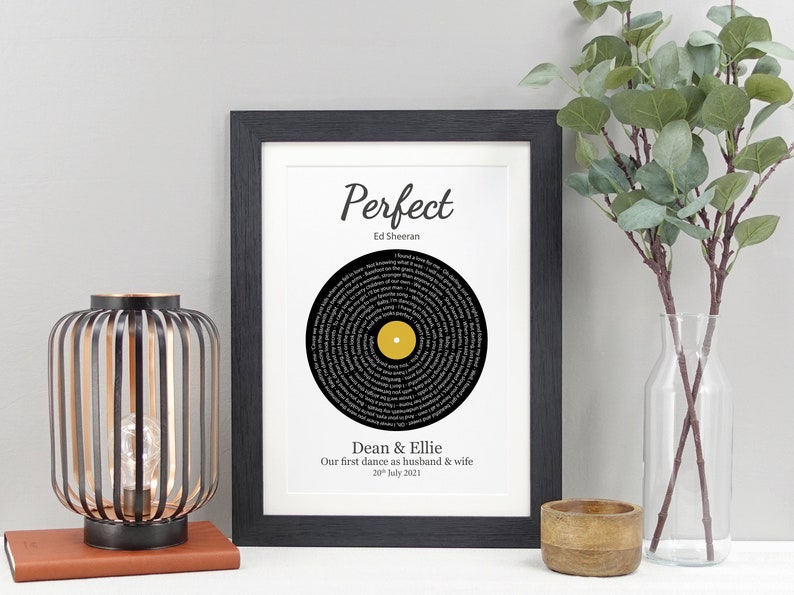 May include: A black framed print with a white background featuring a black vinyl record with the lyrics to the song "Perfect" by Ed Sheeran printed in a circle around the center. The text "Dean & Ellie Our first dance as husband & wife 20th July 2021" is printed below the record.