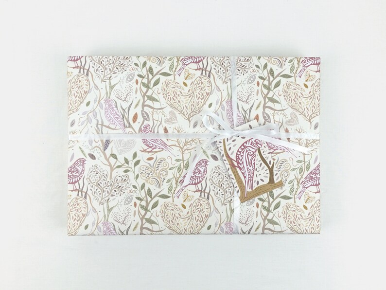 May include: A white gift box with a white ribbon and a brown tag. The box is wrapped in paper with a pattern of birds, leaves, and hearts in shades of pink, brown, and green.