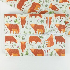 May include: A sheet of wrapping paper with a light green background and a repeating pattern of brown Highland cows with calves. The cows are surrounded by small green and purple flowers.