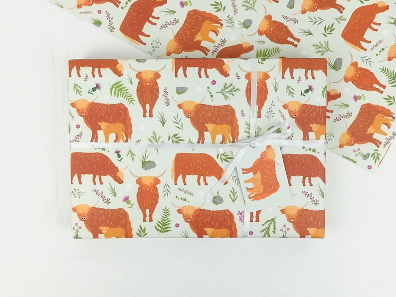 May include: A sheet of wrapping paper with a light green background and a repeating pattern of brown Highland cows with calves. The cows are surrounded by small green and purple flowers.