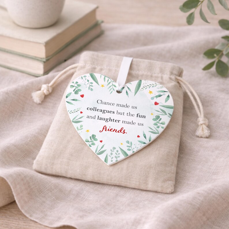 K&ouml;nnte beinhalten: Ein herzf&ouml;rmiger Keramikschmuck mit botanischem Design und den Worten "Chance made us colleagues but the fun and laughter made us friends." Der Schmuck h&auml;ngt an einem beigen Kordelzugbeutel.