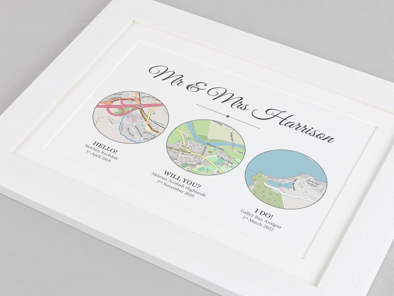 May include: A white framed print with three circular maps representing different locations. The text "Mr & Mrs Harrison" is at the top of the print. The maps are labeled "HELLO! Neschey, Rochdale 1st April 2018", "WILL YOU? Melrose, Scottish Highlands 3rd November 2020", and "I DO! Galley Bay, Antigua 1st March 2022".