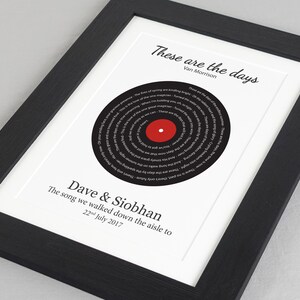 May include: A black and white framed print with a black vinyl record design in the center. The record has the lyrics to the song "These Are the Days" by Van Morrison printed in a spiral pattern. The text "Dave & Siobhan" and "The song we walked down the aisle to 22nd July 2017" is printed below the record.