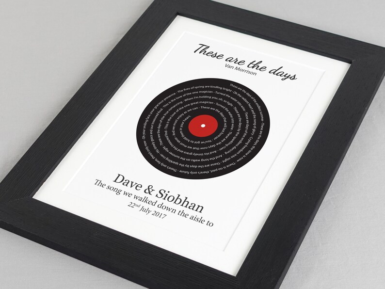 May include: A black and white framed print with a black vinyl record design in the center. The record has the lyrics to the song "These Are the Days" by Van Morrison printed in a spiral pattern. The text "Dave & Siobhan" and "The song we walked down the aisle to 22nd July 2017" is printed below the record.