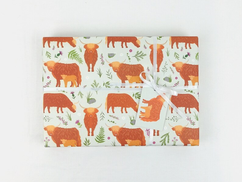 May include: A sheet of wrapping paper with a repeating pattern of brown Highland cows with their calves. The cows are surrounded by green foliage and small pink and purple flowers. The paper is tied with a white ribbon and a gift tag.