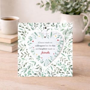K&ouml;nnte beinhalten: Ein wei&szlig;es herzf&ouml;rmiges Ornament mit dem Text "Chance made us colleagues but the fun and laughter made us friends." h&auml;ngt vor einer quadratischen Karte mit gr&uuml;nem Bl&auml;ttermuster. Eine wei&szlig;e Tasse und B&uuml;cher sind im Hintergrund.
