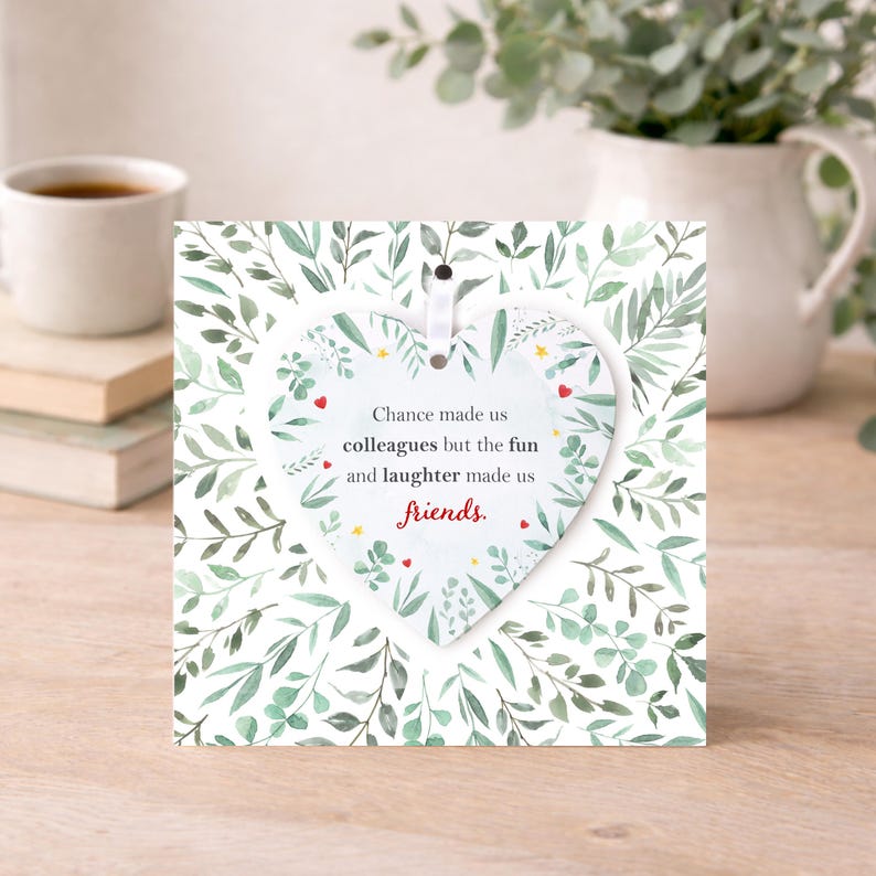 K&ouml;nnte beinhalten: Ein wei&szlig;es herzf&ouml;rmiges Ornament mit dem Text "Chance made us colleagues but the fun and laughter made us friends." h&auml;ngt vor einer quadratischen Karte mit gr&uuml;nem Bl&auml;ttermuster. Eine wei&szlig;e Tasse und B&uuml;cher sind im Hintergrund.