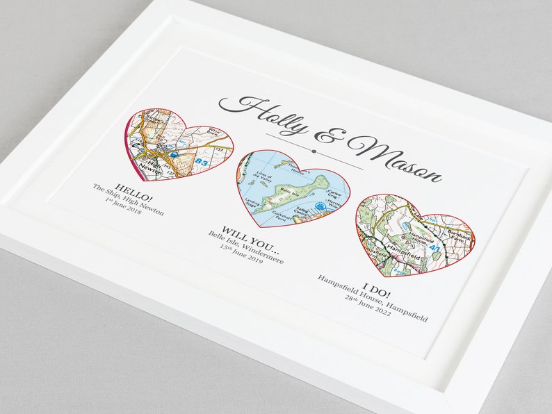 Pu&ograve; includere: Una stampa incorniciata bianca con tre cuori rossi, ognuno contenente una mappa di una posizione diversa. Il testo "Holly & Mason" &egrave; in cima alla stampa. Il testo sotto ogni cuore recita "HELLO! The Ship, High Newton 1 giugno 2018", "WILL YOU... Belle Isle, Windermere 15 giugno 2019", e "I DO! Hampsfield House, Hampsfield 28 giugno 2022".