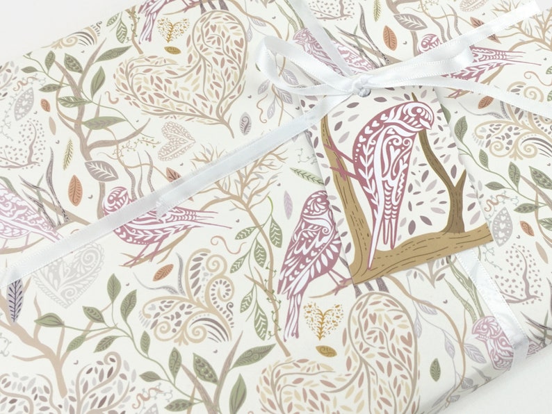 May include: White wrapping paper with a repeating pattern of stylised birds, leaves, and hearts in shades of brown, pink, and green. A white ribbon is tied around the paper with a gift tag featuring a brown branch with a pink bird.