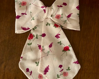May include: A cream-colored bow tie with a floral pattern featuring red, pink, and purple flowers with green leaves. The bow tie has long, pointed ends and is made of a soft, flowing fabric.