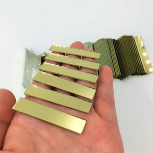 Pode incluir: Uma pilha de azulejos de espelho dourados, cada azulejo é retangular e mede aproximadamente 5 cm de comprimento. Os azulejos estão dispostos em forma de espinha de peixe, com o azulejo superior sendo o mais curto e o azulejo inferior sendo o mais longo.