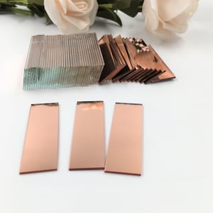 Könnte beinhalten: Ein Satz aus drei rechteckigen roségoldenen Spiegelkacheln. Die Kacheln sind in einer Reihe auf einer weißen Oberfläche angeordnet.