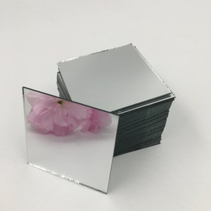 Könnte beinhalten: Ein Stapel kleiner, quadratischer, silberner Spiegel mit einer rosa Blume, die im oberen Spiegel reflektiert wird.