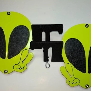 May include: Two bright green alien head-shaped metal signs with black eyes and peace signs. The signs are attached to a black metal stand.