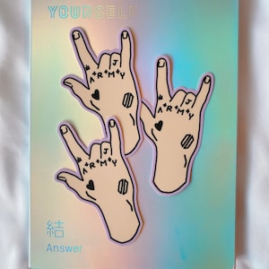 May include: A pink and blue holographic album cover with the text "LOVE YOURSELF" and "Answer" in black. Three stickers of hands making the rock and roll hand gesture are on the cover. The hands have black tattoos that say "*R*M*Y" and "ARMY".
